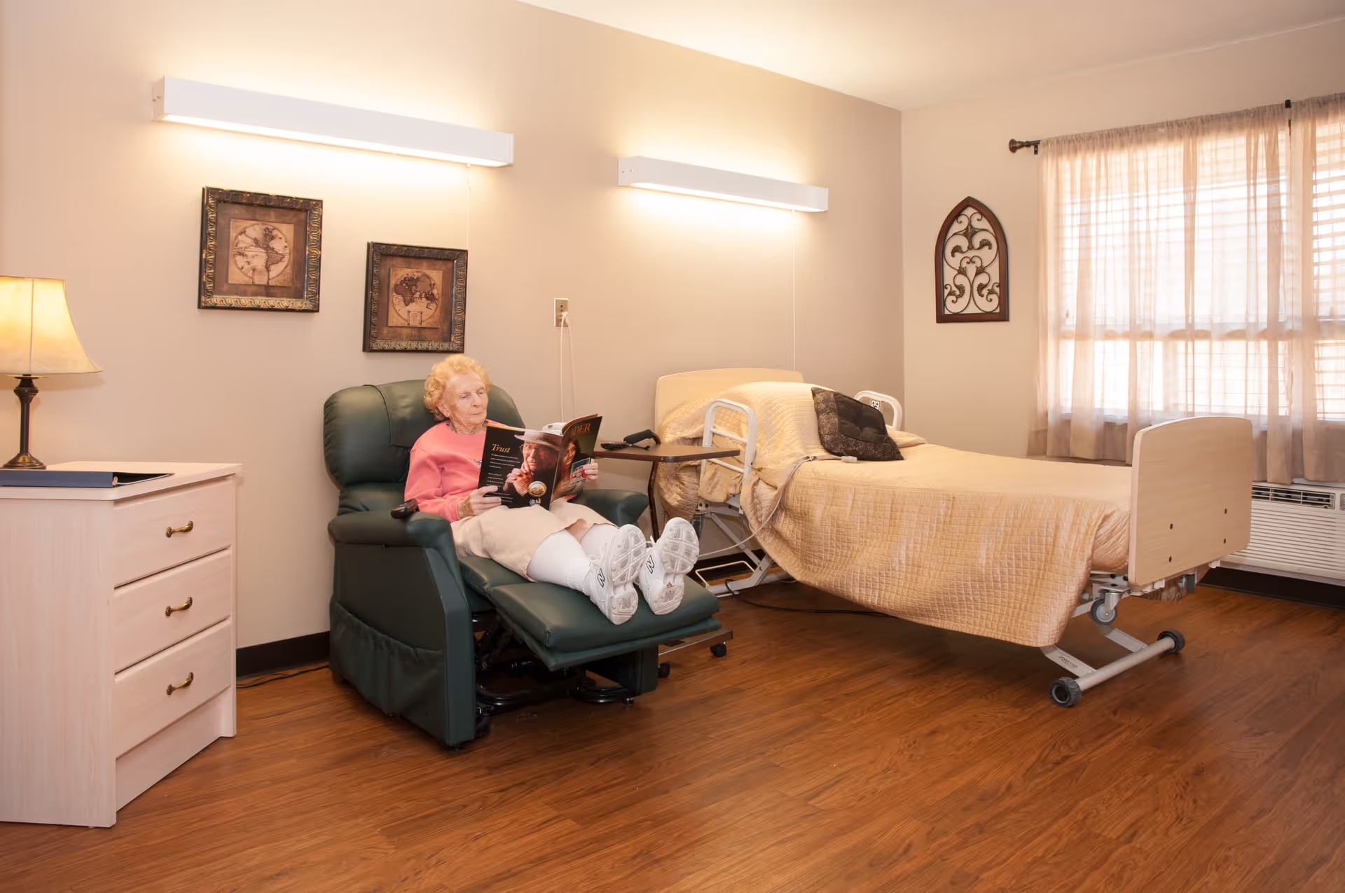 An elderly woman is sitting comfortably in a green recliner chair reading a book in a well-lit room. The room features a hospital-style bed with beige bedding, a wooden nightstand with a lamp, two framed pictures on the wall, a window with sheer curtains, and a decorative wall hanging.