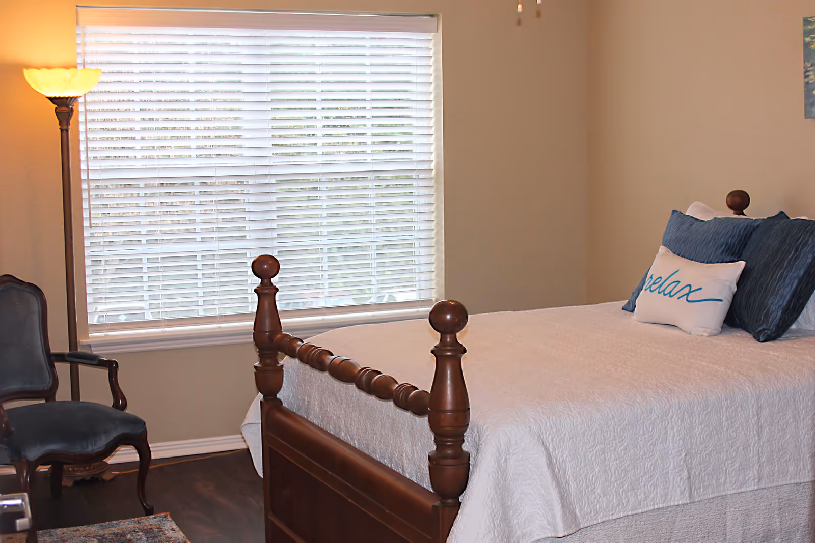 A bedroom with a wooden bed frame, white quilted bedspread, and pillows including one with the word 'relax' written on it. There is a window with white blinds, a floor lamp with a warm light, and a dark upholstered chair next to the bed.