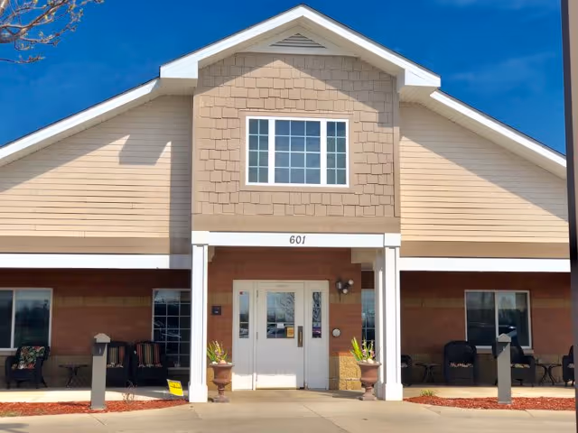 Front exterior view of a senior living facility building with beige siding and a peaked roof. The entrance has double white doors with glass panels, flanked by two columns and two potted plants. There are seating areas with chairs on both sides of the entrance under a covered porch. The building number 601 is displayed above the entrance. The sky is clear and blue.