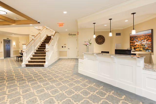 Interior view of a senior living facility lobby featuring a white reception desk with pendant lights above, a large wall clock, a framed landscape picture, a staircase with carpeted steps and wooden handrails, an elevator, and patterned carpet flooring.