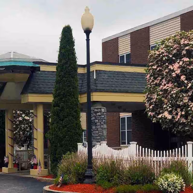 Entrance area of Orchard View Manor featuring a covered driveway with yellow pillars, a tall lamppost, a tall evergreen tree, a flowering tree with pink blossoms, and a white picket fence surrounding landscaped bushes and plants with red mulch.