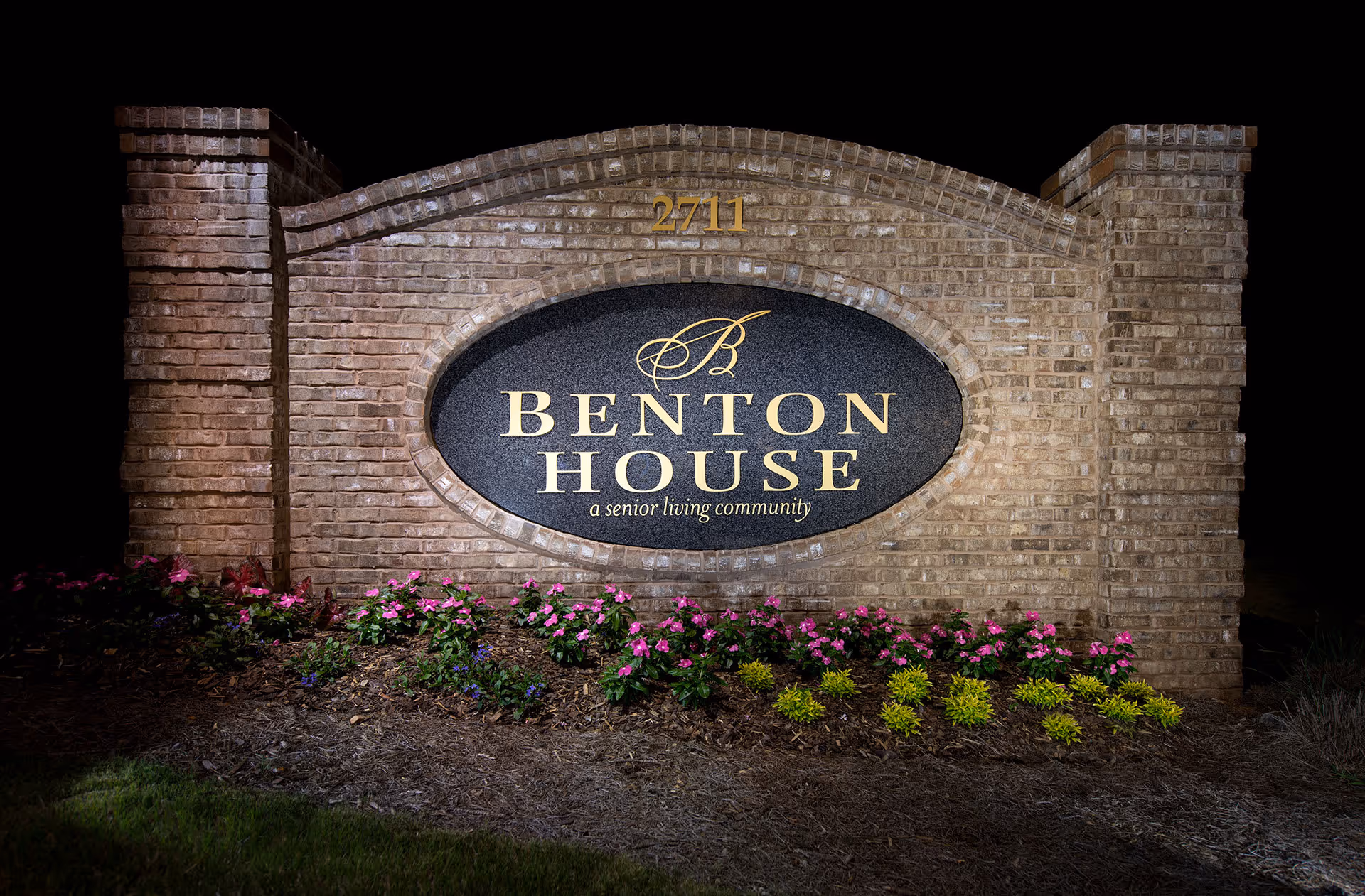 Illuminated brick entrance sign reading "Benton House, a senior living community" with flowers planted at its base.