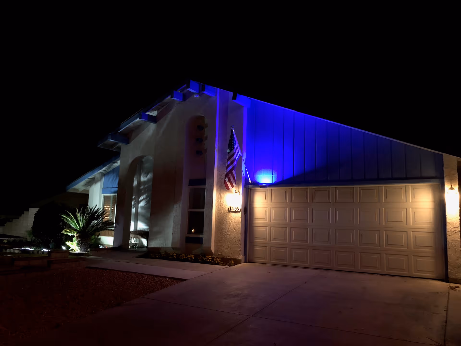 Night view of the front of a single-family home with a lit garage, American flag, and blue uplighting.