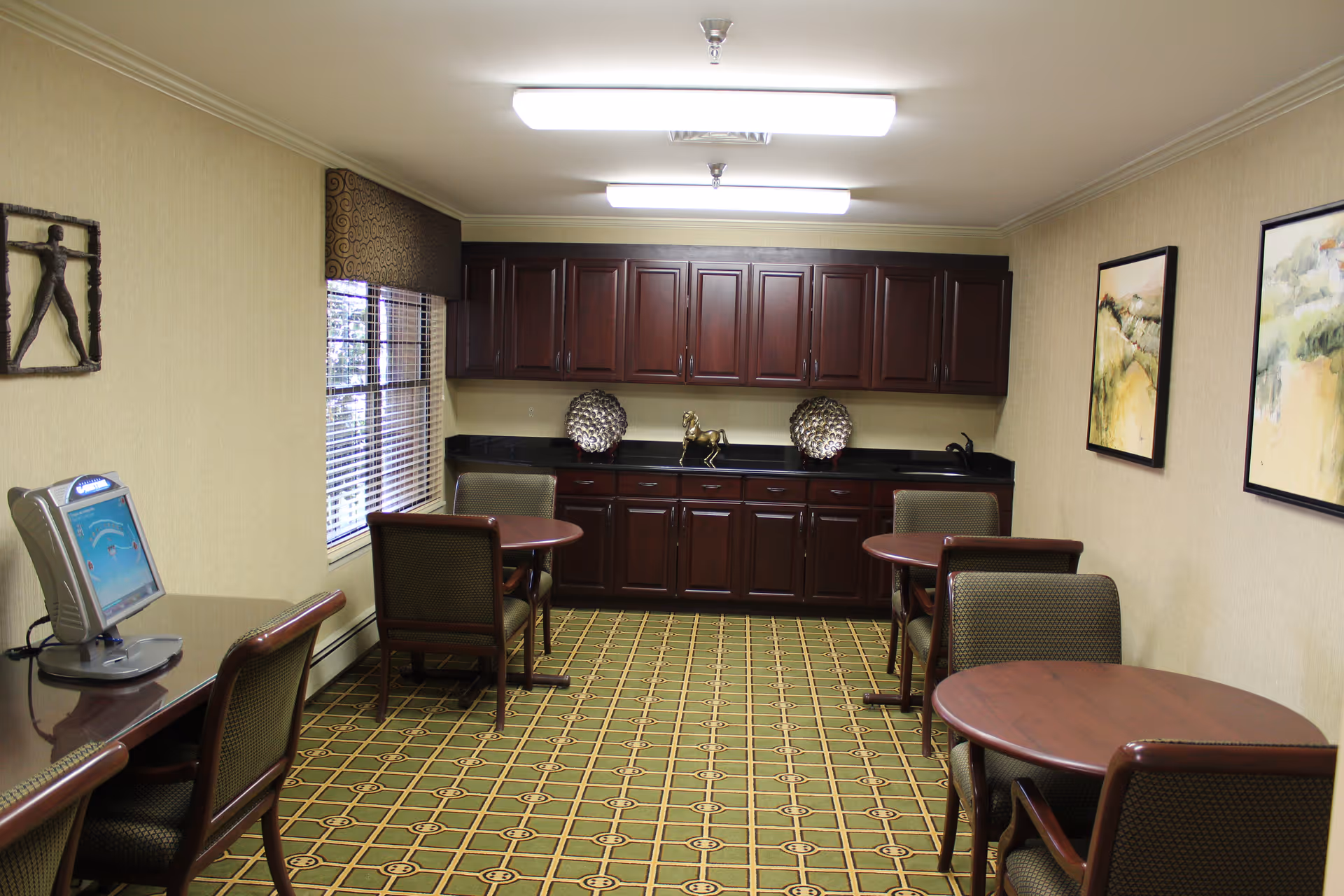 A small room with green patterned carpet, beige walls, and fluorescent ceiling lights. The room contains three round wooden tables each with two upholstered chairs. On the left side, there is a long wooden desk with a computer monitor. At the far end, there are dark wooden cabinets with decorative items on the countertop. Two framed paintings hang on the right wall, and a window with blinds is on the left wall.