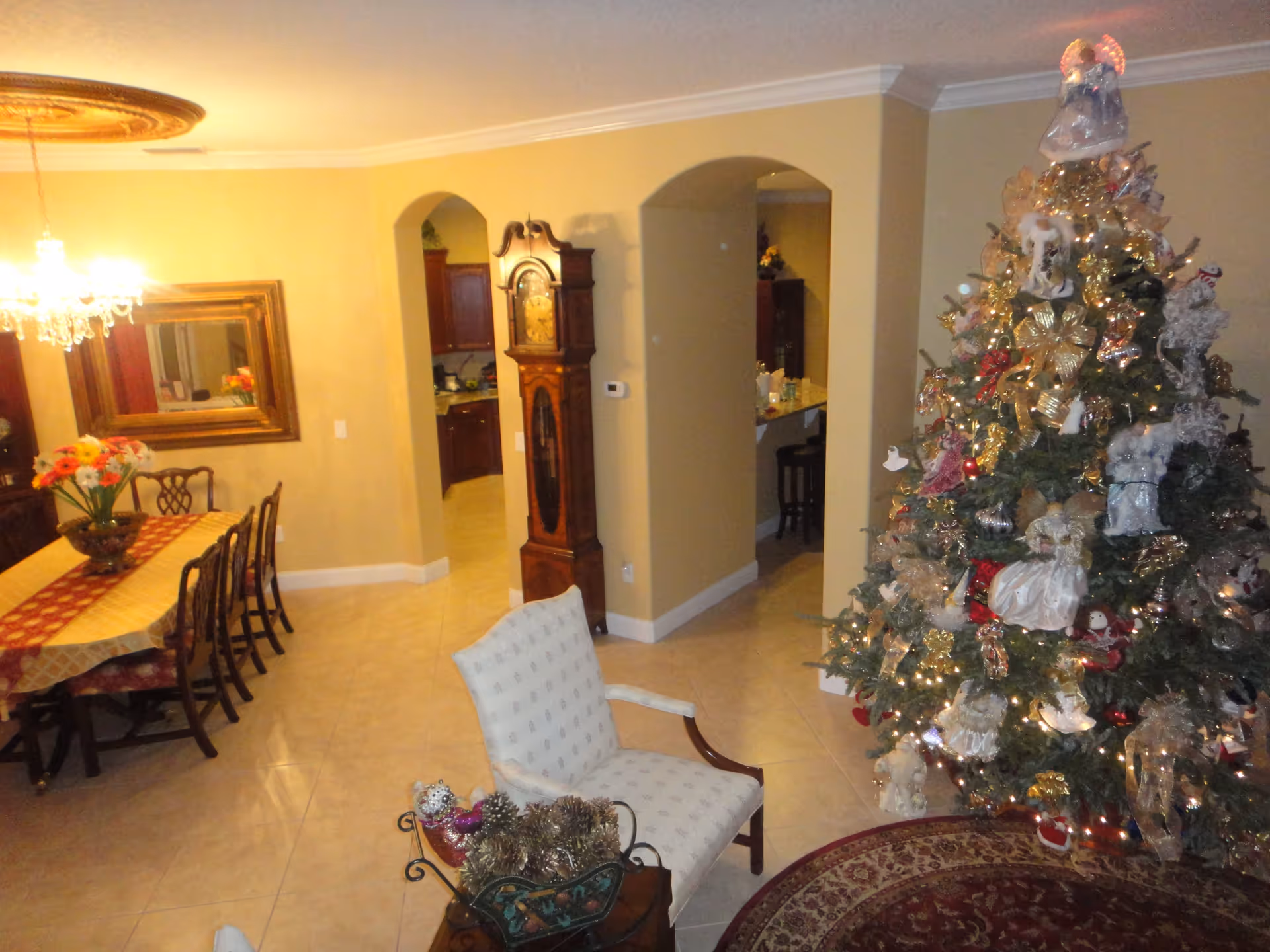 Interior view of a home decorated for Christmas with a large Christmas tree adorned with lights, ribbons, and ornaments. To the left, there is a dining area with a table covered by a tablecloth and surrounded by chairs, a chandelier hanging above, and a large mirror on the wall. In the center, there is a grandfather clock and an upholstered armchair with a small table holding pinecones and decorative items. The walls are painted beige, and the floor is tiled.