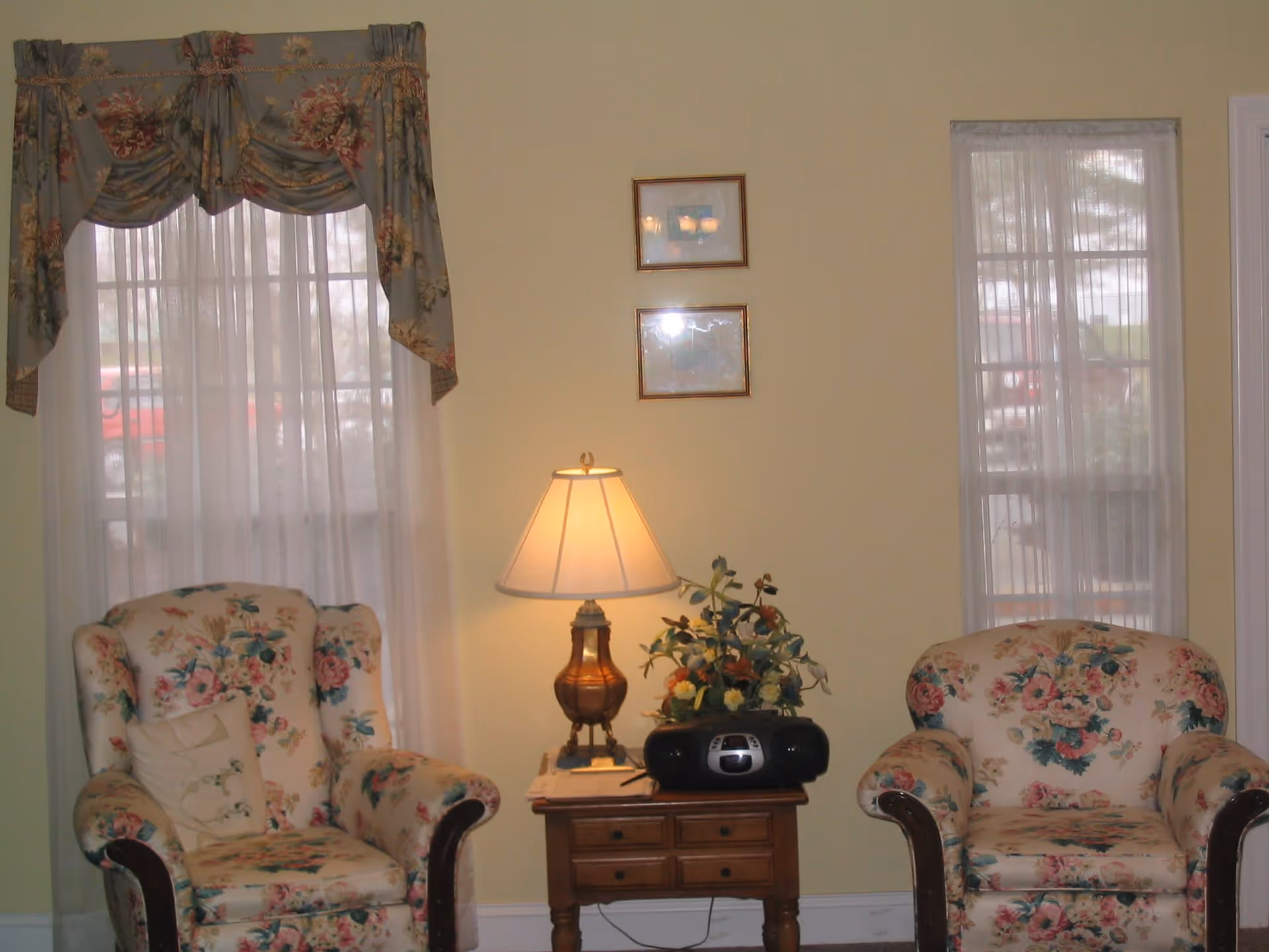 A cozy sitting area with two floral upholstered armchairs placed on either side of a wooden side table. The table holds a decorative lamp with a beige shade, a floral arrangement, and a black radio or CD player. Behind the chairs are two windows with sheer white curtains and floral valances. The walls are painted light yellow and have two small framed pictures hanging between the windows.