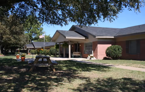Exterior view of a single-story brick building with a covered entrance and a bench outside. There is a picnic table on the grassy lawn in front of the building, with trees providing shade. An American flag is visible near the building.