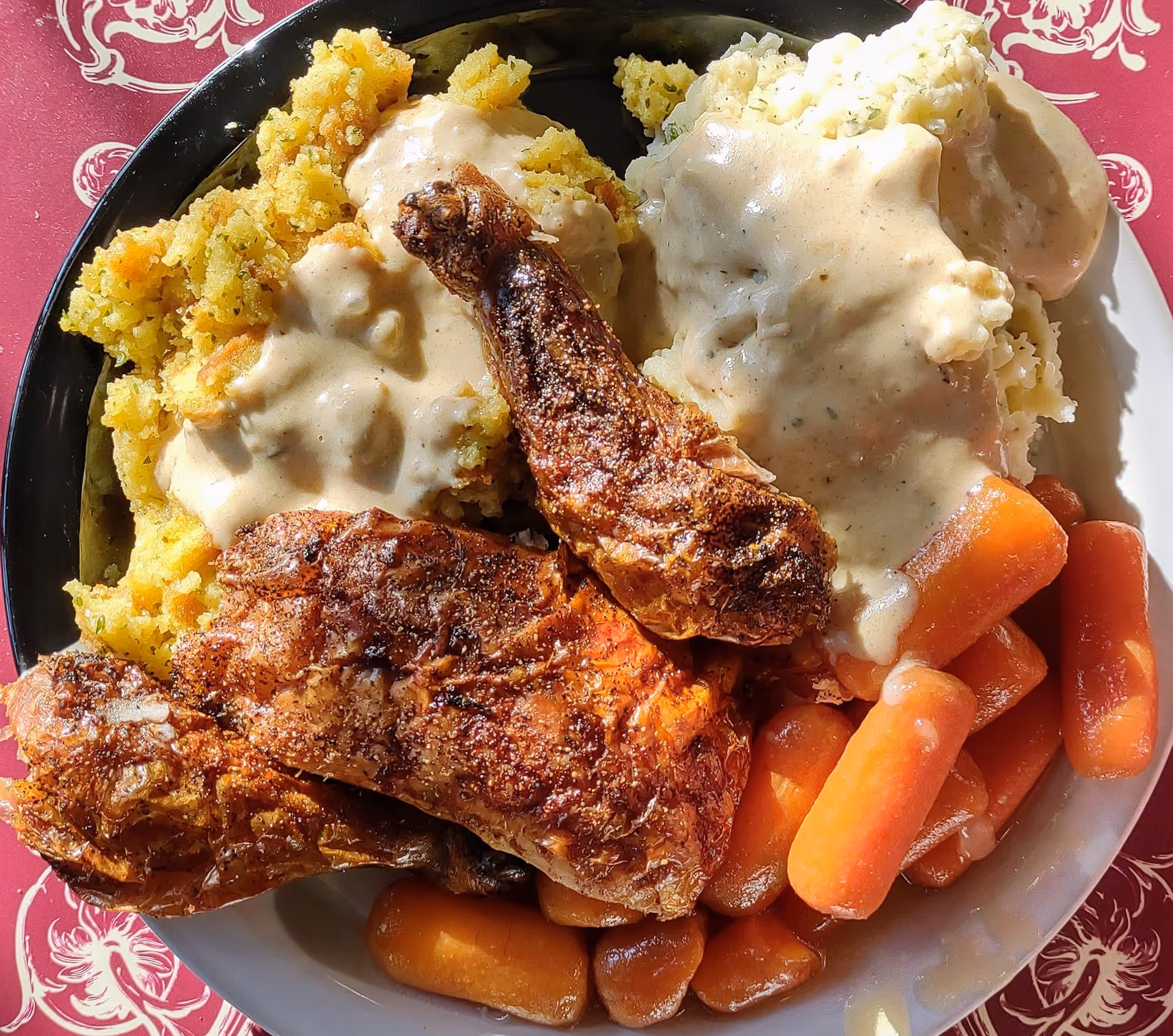 A plate of food featuring crispy fried chicken wings, mashed potatoes with gravy, stuffing with gravy, and cooked baby carrots on a white plate with a red patterned tablecloth underneath.
