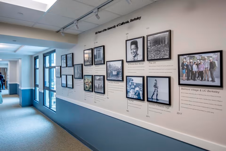 A hallway in Belmont Village Senior Living Albany with a timeline display on the wall titled '150 Years of the University of California, Berkeley,' featuring framed historical photos and descriptions. The hallway has windows on one side and carpeted flooring.