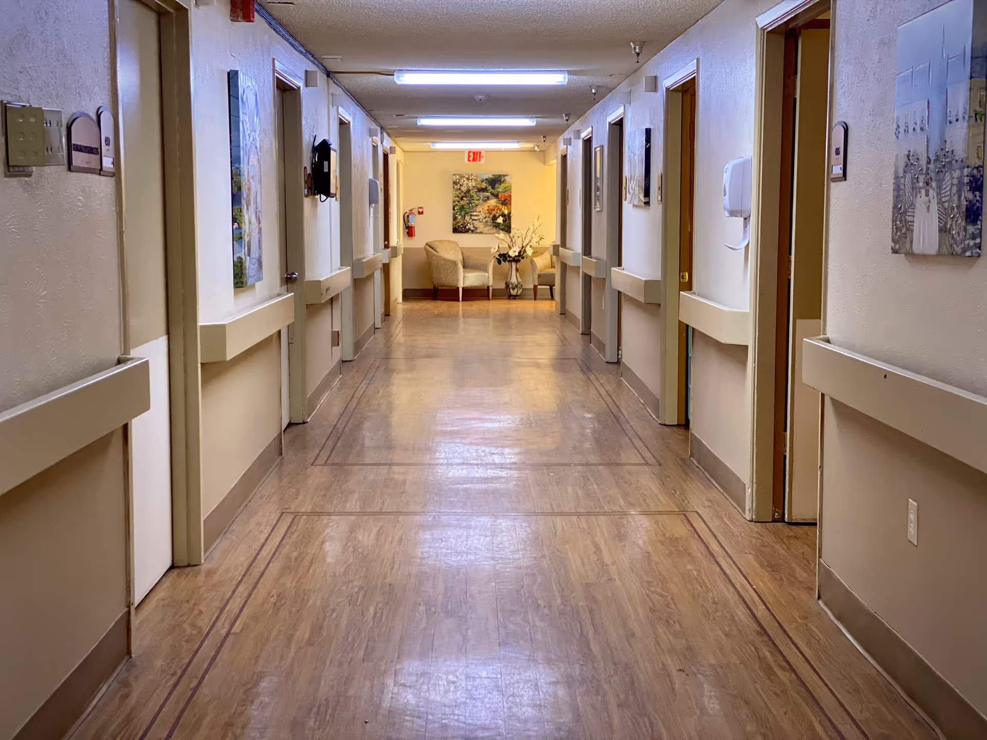 A long, well-lit hallway in a healthcare facility with multiple closed doors on both sides. The floor has a wood-like finish, and the walls are painted in light colors with handrails along both sides. At the end of the hallway, there are two beige armchairs and a small table with a flower arrangement, beneath a colorful painting. Ceiling lights illuminate the corridor, and an exit sign is visible above the seating area.