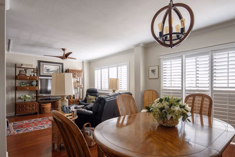 Open-plan living and dining area with a wooden dining table and chairs with a floral centerpiece, leather sofa, TV shelving, and shuttered windows.