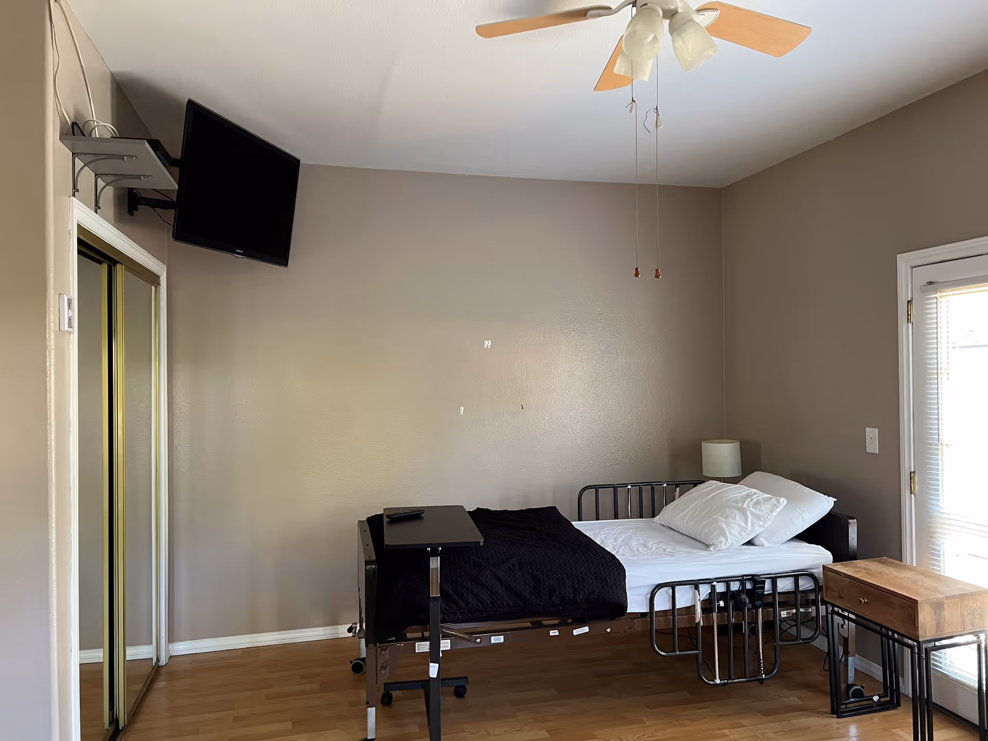 A simple bedroom with a single hospital-style bed with white sheets and pillows, a black blanket, a small wooden side table, a wall-mounted TV, a ceiling fan with light, and a mirrored closet door. The walls are painted beige and there is a door with blinds letting in natural light.