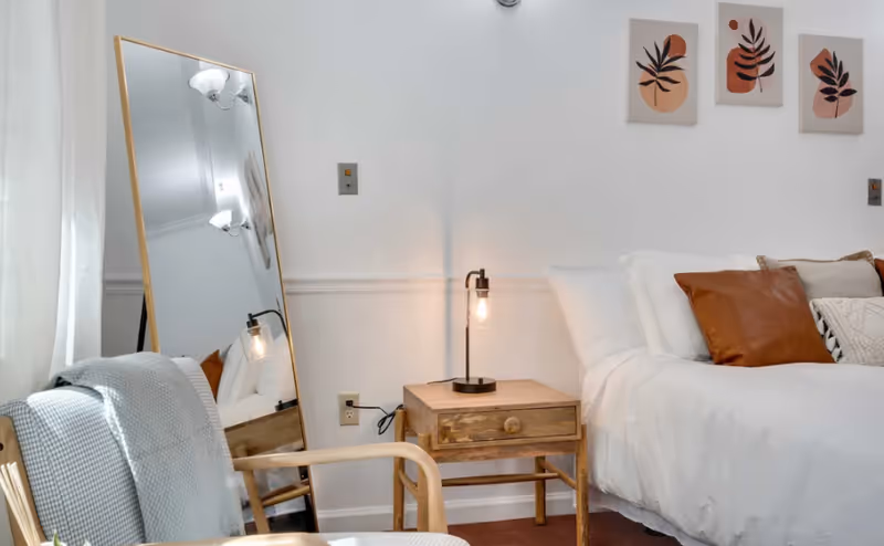 Bright bedroom featuring a white bed with brown pillows, a wooden nightstand and lamp, a leaning full-length mirror, and a chair draped with a blanket.