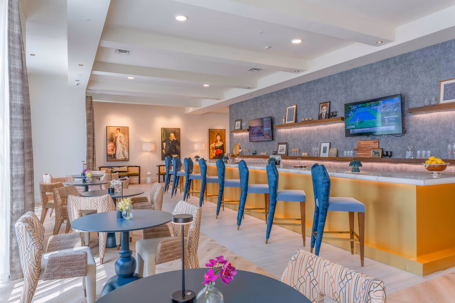 A modern and stylish dining area with round tables and patterned chairs. There is a long bar with blue high chairs and a yellow base. The back wall features two mounted flat-screen TVs, shelves with framed pictures, and decorative items. Three portrait paintings hang on the far wall, and the space is well-lit with natural light from large windows covered with sheer curtains.