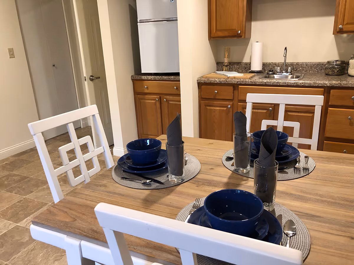 A dining table set for three with blue bowls, plates, and silverware on round placemats. Each place setting has a glass with a folded dark napkin. The table is in a kitchen area with wooden cabinets, a countertop with a sink, and a refrigerator in the background.