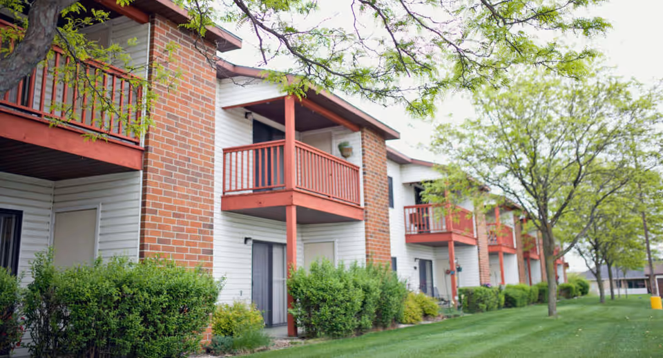 Exterior view of a senior living facility named The Meadows showing a row of two-story buildings with white siding and red brick chimneys. Each unit has a red wooden balcony or patio, surrounded by green bushes and trees with fresh green leaves. The grass is well-maintained and the sky is overcast.
