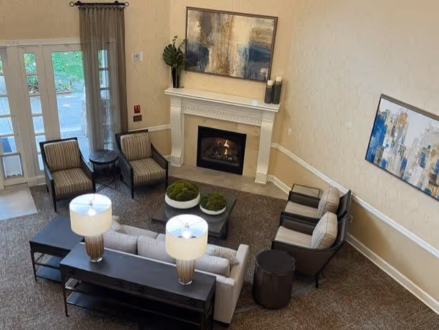 A cozy senior living facility common area with a fireplace, two table lamps on a console table behind a sofa, four armchairs arranged around a coffee table with decorative moss balls, and two abstract paintings on the walls. There is a glass door with curtains leading outside.