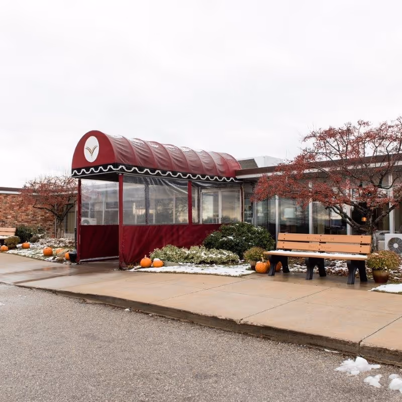 Front entrance of a senior living facility with a maroon covered canopy, bench, pumpkins, and light snow on the ground.