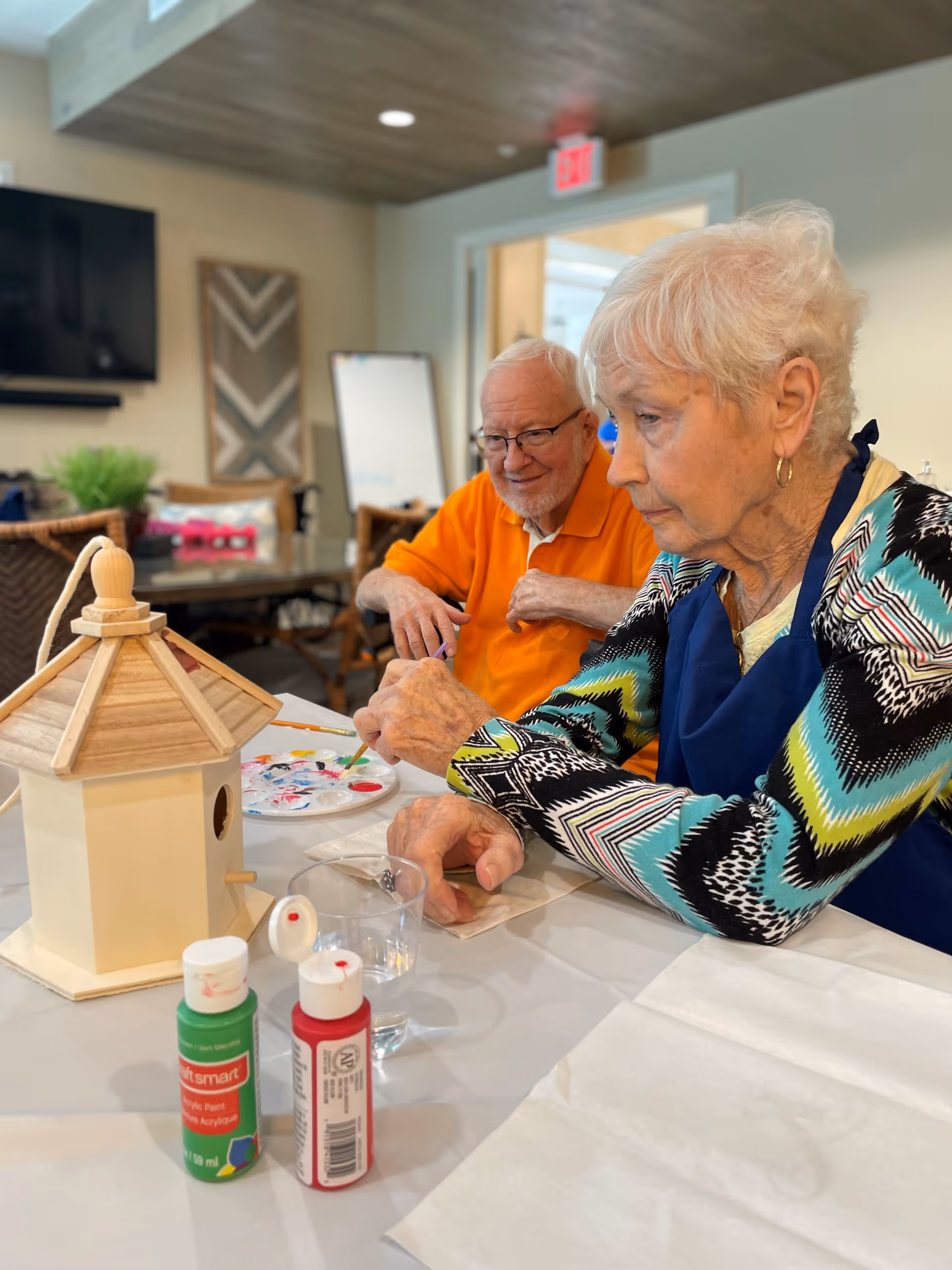 An elderly woman and man sitting at a table indoors, painting a wooden birdhouse. The woman is focused on painting while the man looks on with a smile. There are paint bottles, a paint palette, and a glass of water on the table. The room has a cozy atmosphere with a TV, wall art, and wicker chairs in the background.