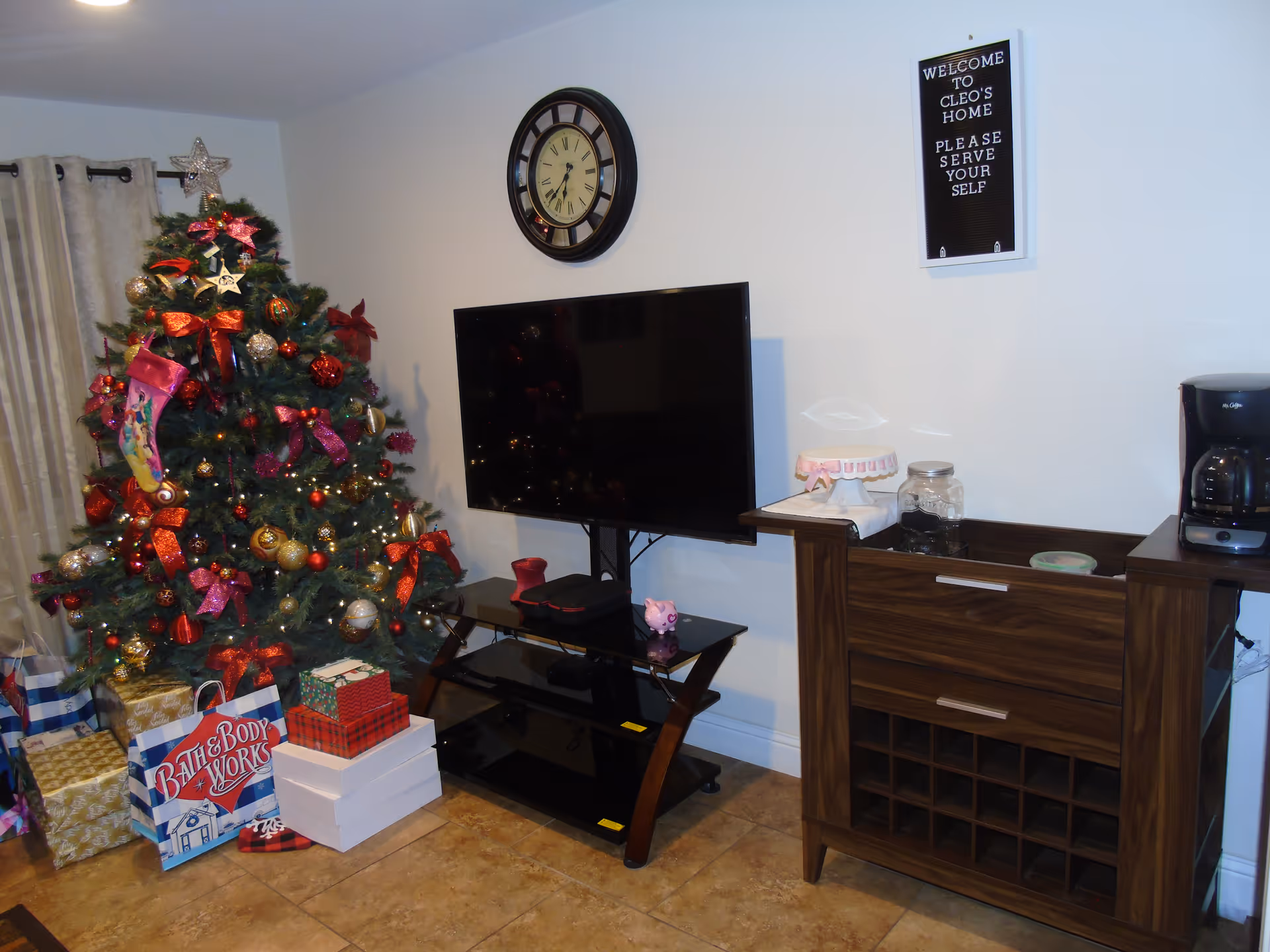 Decorated living room with a Christmas tree, wrapped presents, flat-screen TV, wall clock, and a wooden cabinet with a sign reading "Welcome to Cleo's Home."