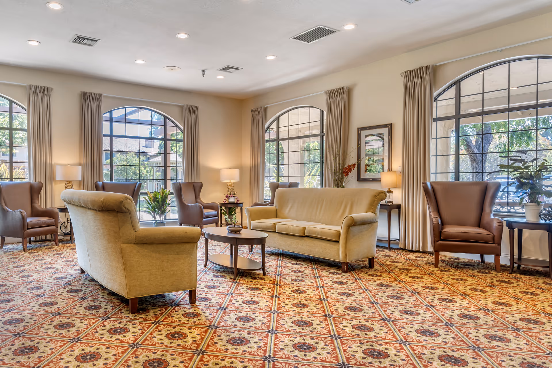 A spacious living room area with large arched windows letting in natural light. The room features beige sofas, brown armchairs, small tables with lamps, and decorative plants. The floor is covered with a patterned carpet in warm tones.