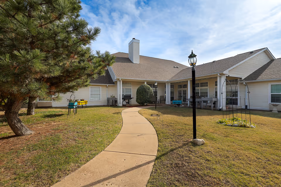 Outdoor view of a senior living facility with a curved concrete pathway leading to a building with beige siding and a brown roof. The area features a large tree on the left, a lamp post in the center, garden planters, and a small garden bed with a metal arch on the right under a partly cloudy sky.