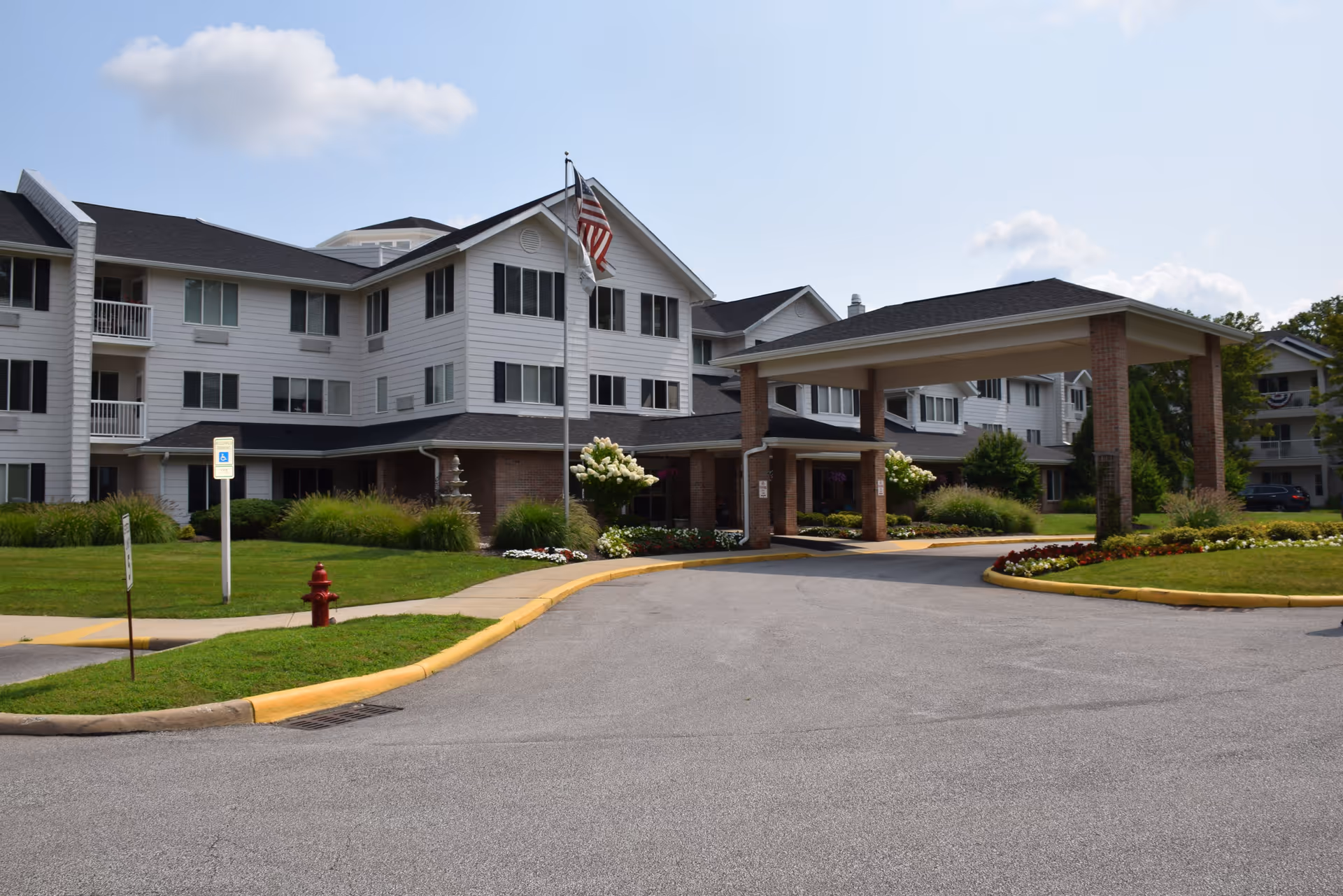 Exterior view of a multi-story senior living facility with white siding and black shutters. The building has a covered entrance with brick pillars, a driveway, and landscaped greenery including flowers and bushes. An American flag is flying on a flagpole near the entrance.