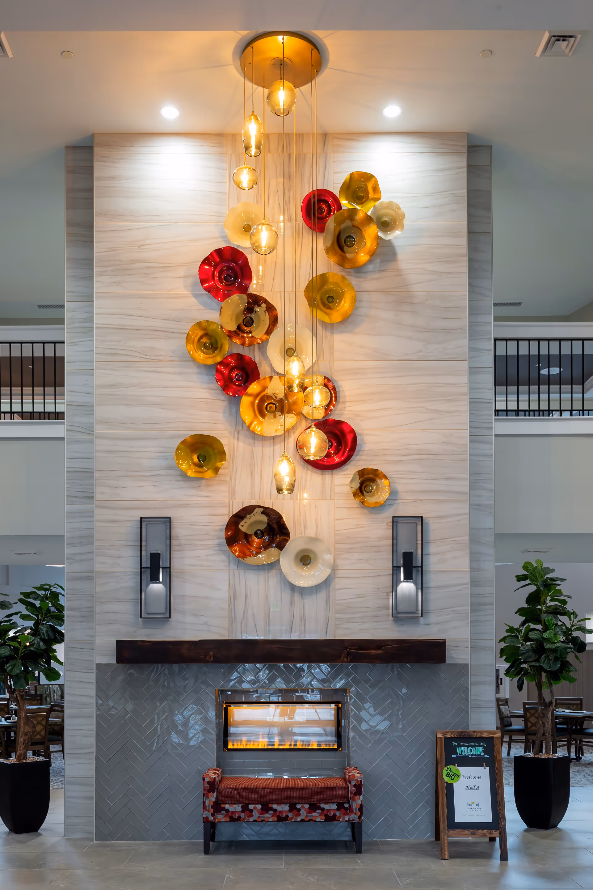 A modern indoor fireplace with a decorative wall featuring colorful glass art pieces in red, yellow, and cream colors. Above the fireplace, several hanging pendant lights with warm bulbs illuminate the area. There is a small upholstered bench with a patterned fabric in front of the fireplace. On the right side, a wooden framed signboard welcomes visitors. Two large potted plants flank the fireplace on either side, and the background shows a spacious interior with tables and chairs.