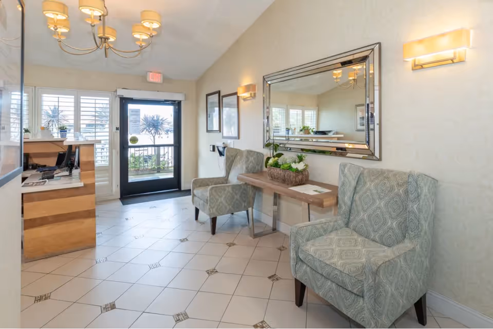 A bright and welcoming reception area with two patterned armchairs, a wooden console table with a flower arrangement, a large decorative mirror on the wall, and a wooden reception desk near the entrance door with windows featuring white shutters.