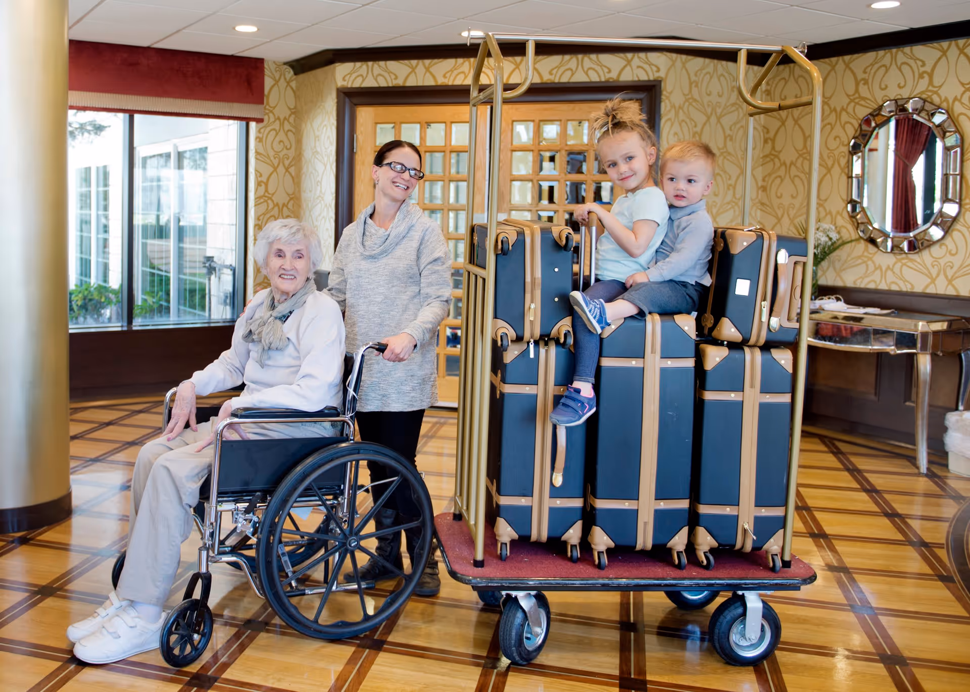 An elderly woman in a wheelchair, a caregiver, and two children sitting on a luggage cart in a decorated lobby.