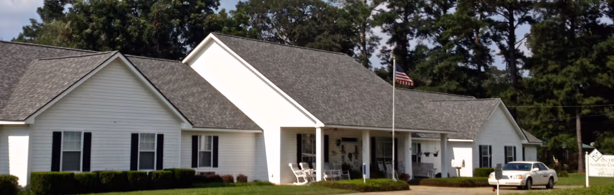 Front exterior of a single-story white building with a covered porch, American flagpole, parked car, and lawn.