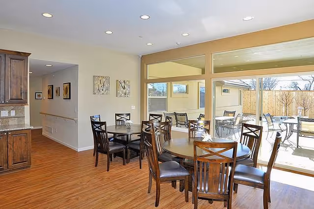 Interior view of a dining area in an assisted living facility with round wooden tables and chairs. The room has large windows and sliding glass doors that open to an outdoor patio with additional seating. The floor is wood, and the walls are painted light beige. There are framed artworks on the wall and recessed ceiling lights.