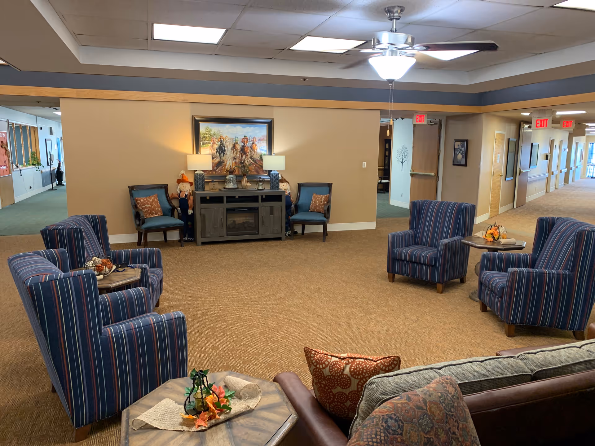 A cozy common area in an assisted living facility with several striped armchairs arranged around small tables decorated with autumn-themed centerpieces. There is a brown couch with patterned pillows in the foreground. Against the far wall is a console table with two lamps and decorative items, flanked by two blue chairs and two scarecrow decorations. A painting of horse racing hangs above the console table. The ceiling has a fan with a light, and the hallway with exit signs is visible in the background.