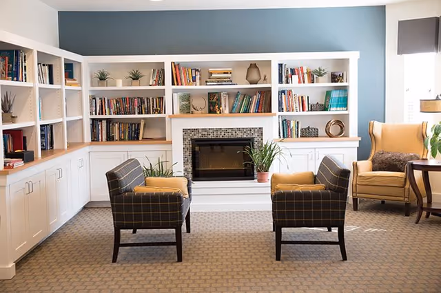 A cozy senior living common area with two plaid armchairs facing a fireplace surrounded by built-in white bookshelves filled with books and decorative items. There is a yellow armchair with a brown cushion near a window with a side table and lamp. The walls are painted blue above the white shelves.