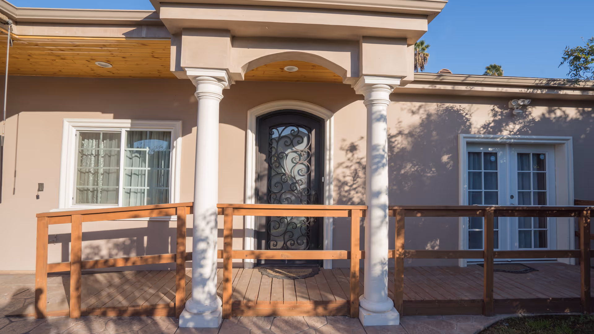 Exterior view of a senior living facility entrance with a decorative black front door framed by two white columns, a wooden ramp with railings, and windows on either side under a clear blue sky.