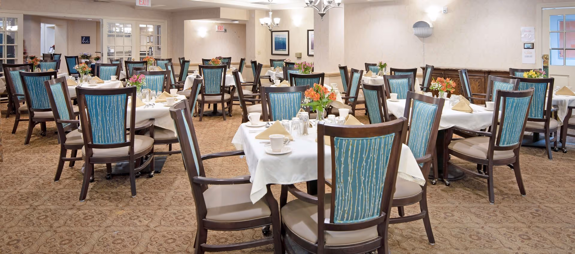 A dining room with multiple tables covered in white tablecloths, each set with cups, glasses, napkins, and floral centerpieces. The chairs have wooden frames with blue patterned upholstery. The room has beige walls, carpeted floors, and soft lighting from wall sconces and chandeliers. There are exit signs and framed artwork on the walls.