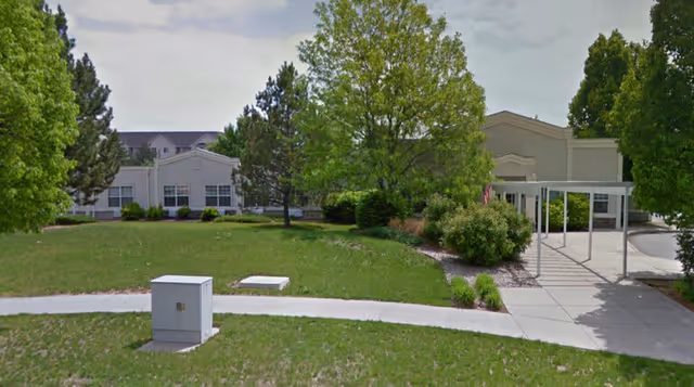 Exterior view of a senior living facility with a green lawn, trees, and a paved walkway leading to the building entrance under a covered walkway.