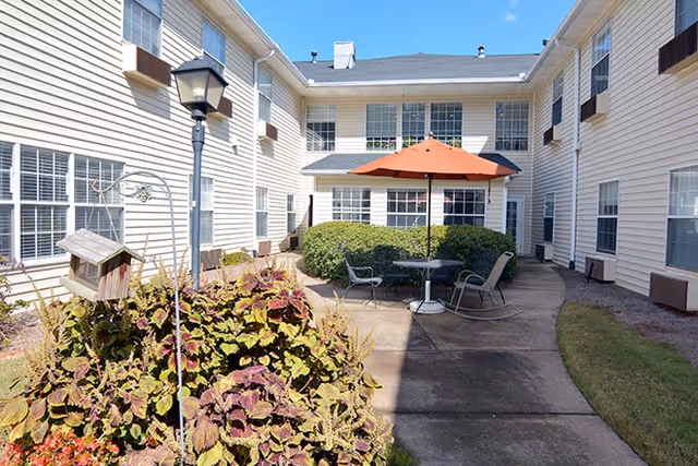 Sunny courtyard patio with table, chairs and an orange umbrella surrounded by a two-story beige siding senior living building.