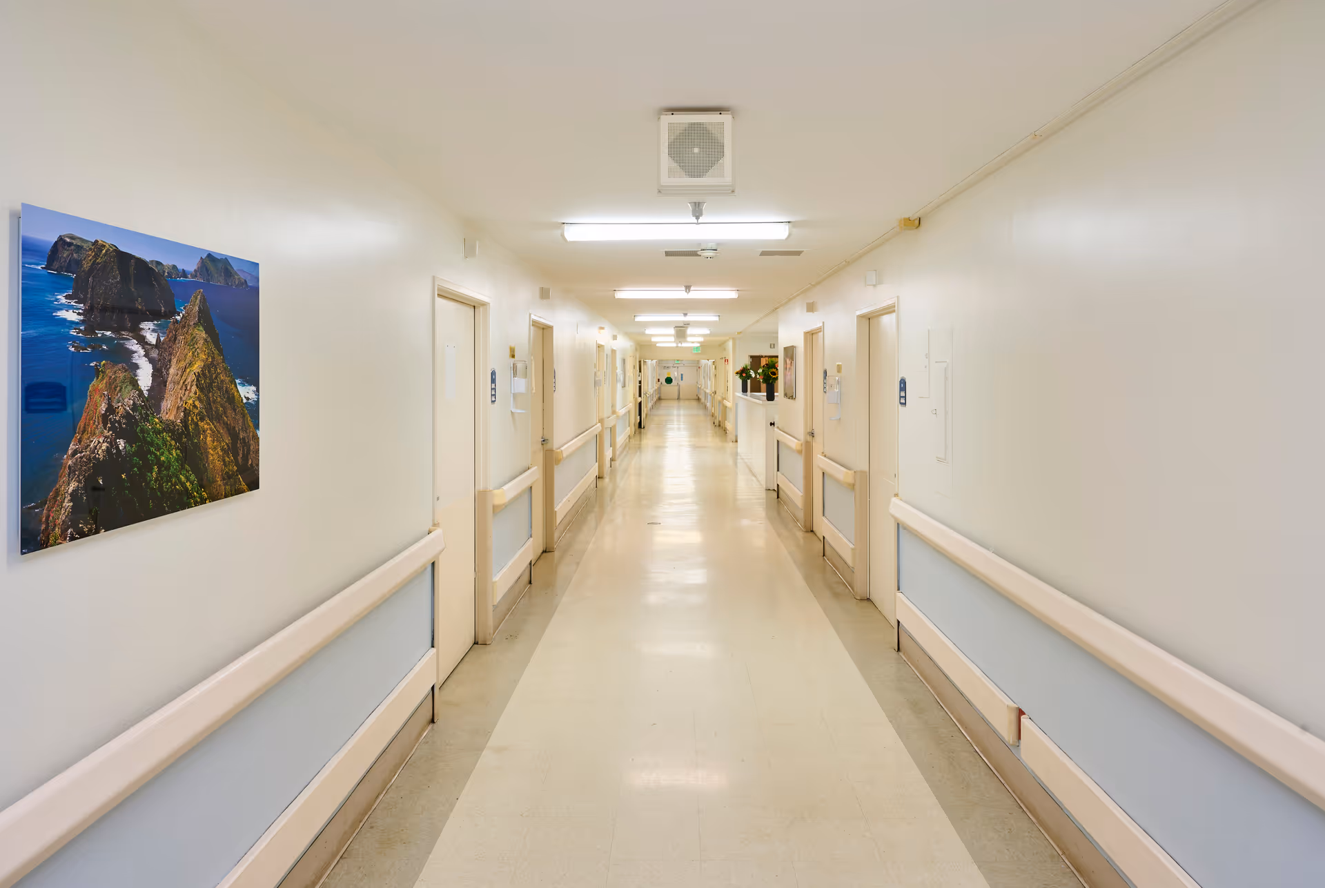 A long, clean hallway in a care facility with white walls, beige handrails, and several closed doors on both sides. The ceiling has fluorescent lights, and there is a large photograph of a rocky coastline hanging on the left wall.
