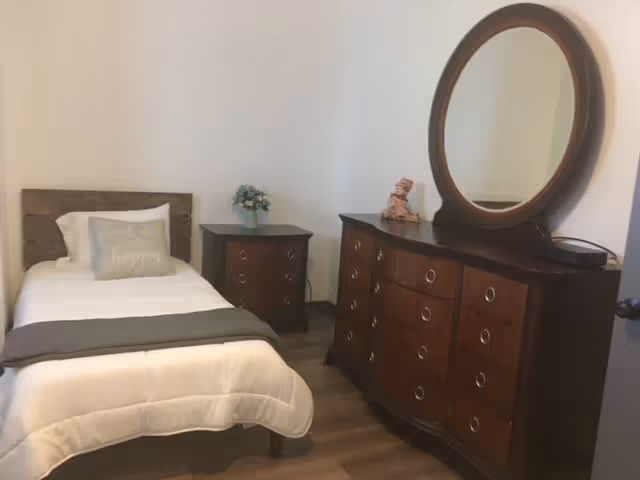 A small bedroom with a single bed featuring white bedding and a gray throw blanket. There is a wooden nightstand with a small flower arrangement next to the bed. Across from the bed is a large wooden dresser with multiple drawers and a round mirror on top. The room has light-colored walls and wooden flooring.