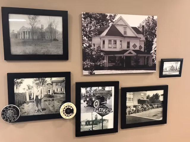 A beige wall with six framed black-and-white photographs depicting historic buildings, a vintage car, and a man standing outdoors. The photos are arranged in a collage style with two circular decorative objects attached to the bottom left photo.