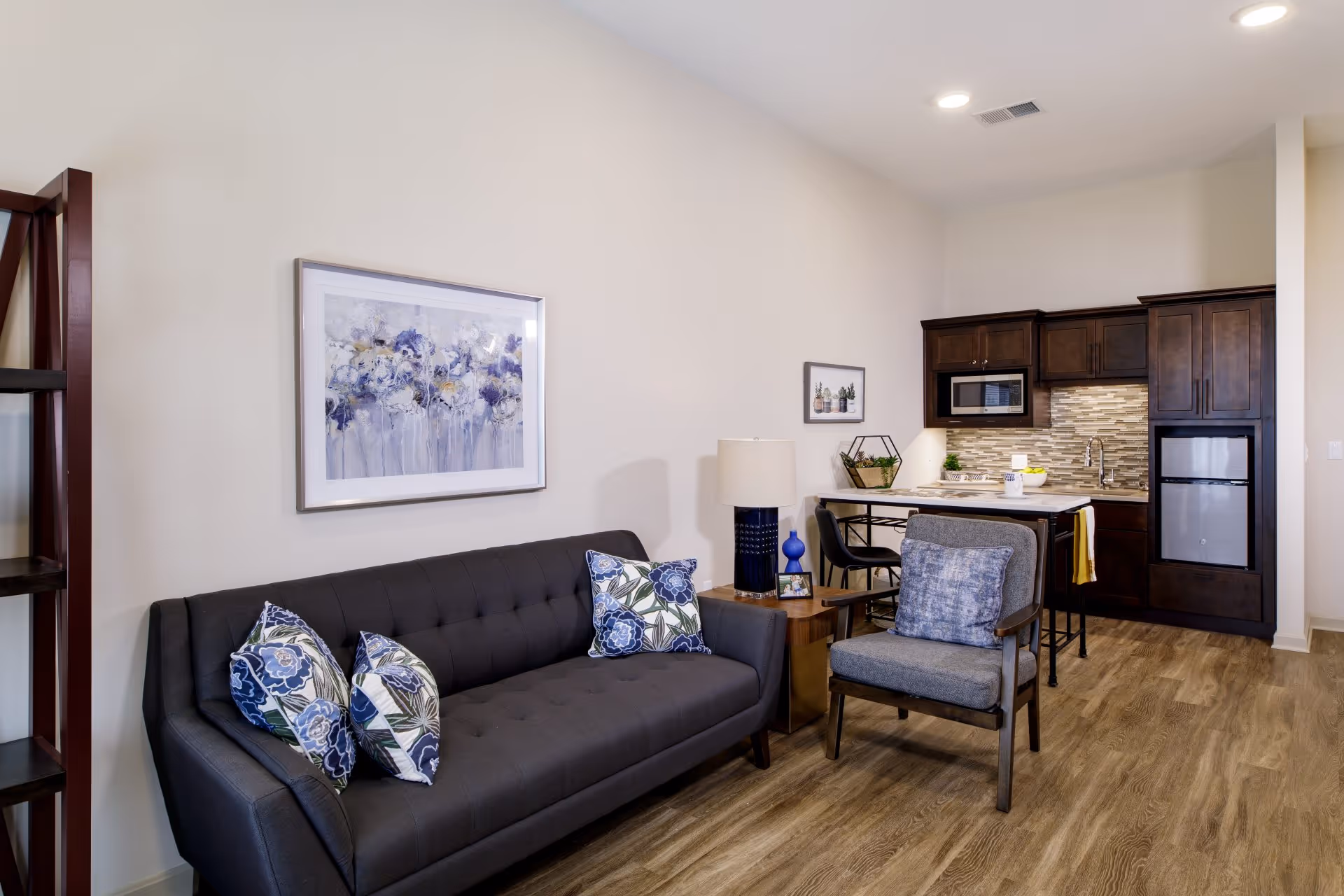 A modern living area with a dark gray sofa adorned with floral patterned pillows, a gray cushioned armchair with a blue pillow, a wooden side table with a blue lamp, and a small kitchen in the background featuring dark wood cabinets, a microwave, a mini refrigerator, and a tiled backsplash.