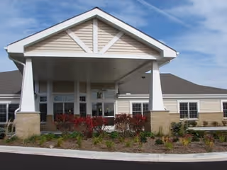 Front exterior view of a single-story building with a covered entrance supported by white pillars, beige siding, and a landscaped area with shrubs and plants in front.