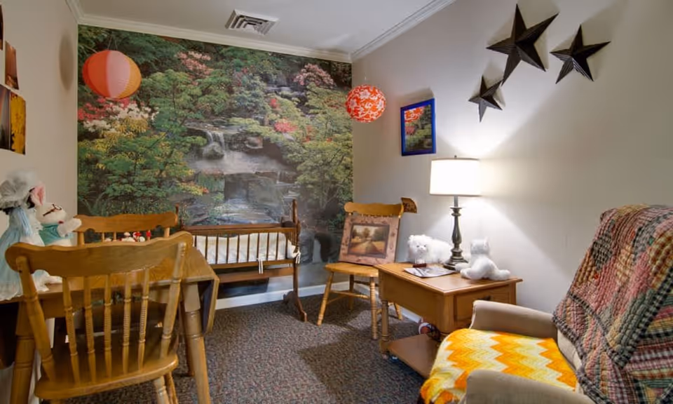 A cozy room with wooden chairs and a small wooden table, decorated with stuffed animals and a lamp. The back wall features a large mural of a serene garden with trees and a waterfall. The room has a carpeted floor and is decorated with hanging paper lanterns and star-shaped wall decorations.