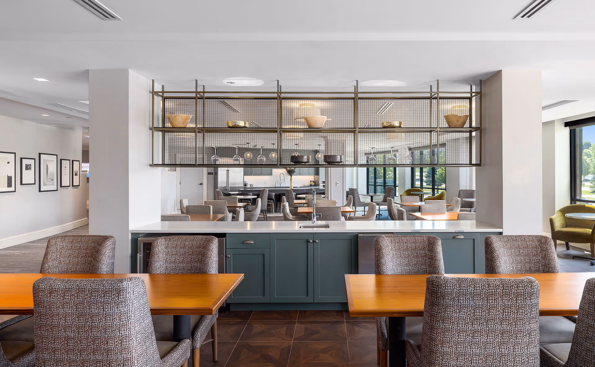Modern dining area with wooden tables and upholstered chairs, a green cabinet with a white countertop and sink in the foreground, and a metal shelving unit with decorative bowls and hanging wine glasses. Large windows allow natural light to fill the space.