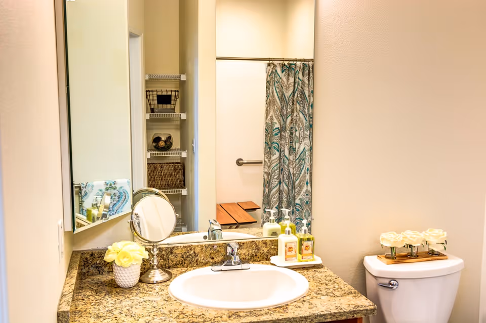 A bathroom with a granite countertop sink, a small round mirror, a vase with yellow flowers, and three bottles of soap or lotion on a tray. In the background, there is a toilet with a wooden tray holding white flowers and a shower with a patterned curtain and a grab bar. Shelves with baskets are visible through the doorway.