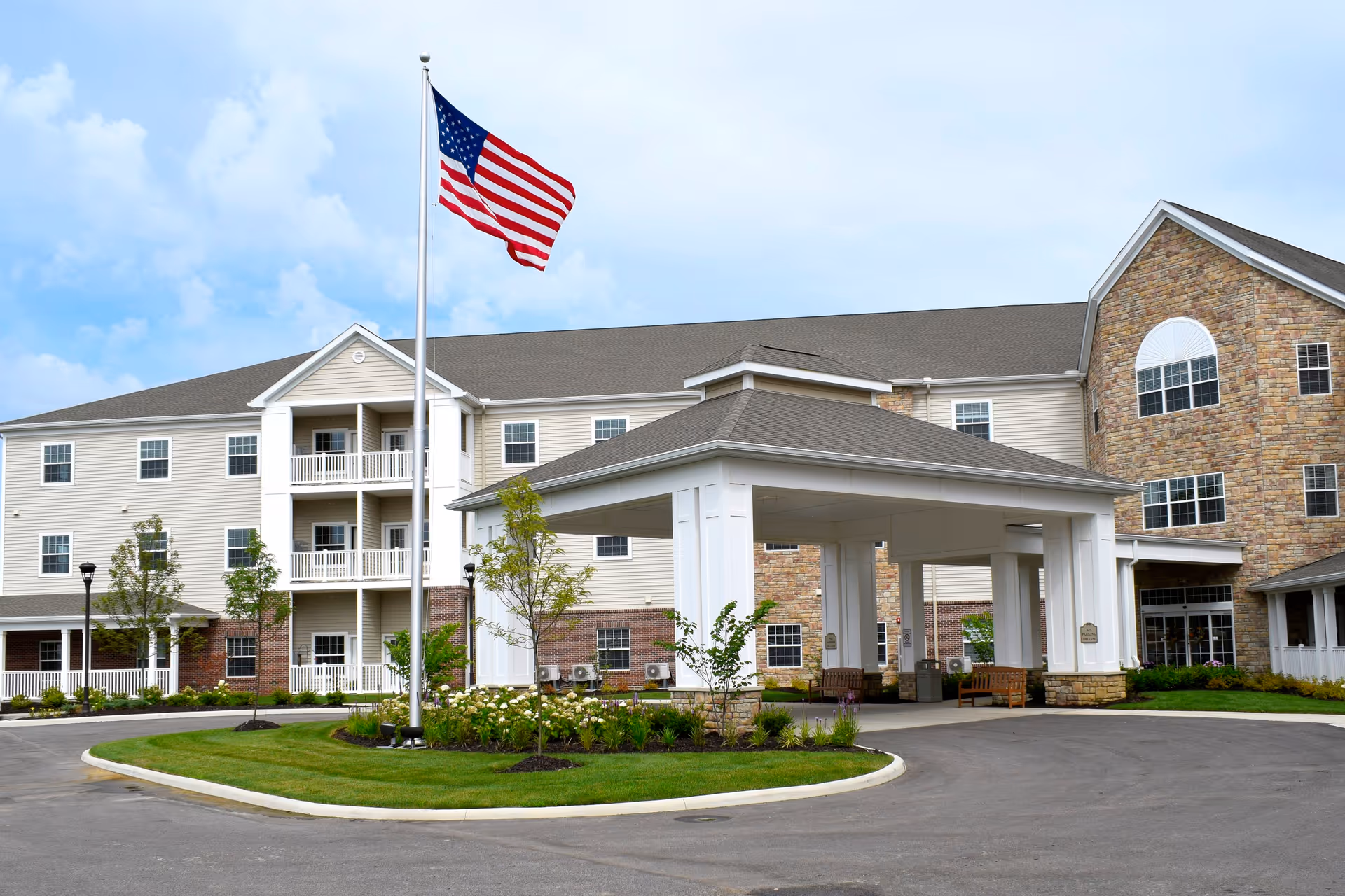 Exterior view of Danbury Hudson senior living facility showing a large building with beige siding and stone accents, a covered entrance with white pillars, a circular driveway, landscaped greenery, and an American flag on a flagpole against a partly cloudy sky.