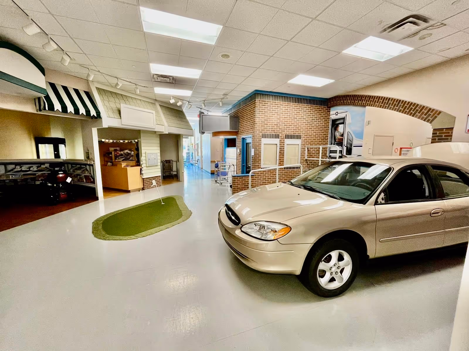 Indoor area resembling a small town street scene with a beige car parked on the right, a miniature putting green in the center, and building facades with brick and siding details under fluorescent ceiling lights.