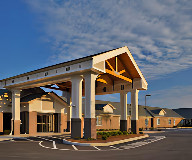 Exterior view of Harbour Manor & The Lodge building entrance with a large covered drop-off area supported by white columns, under a partly cloudy blue sky.