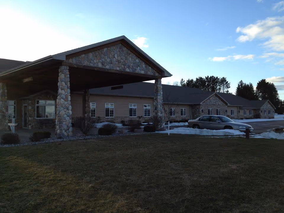 Single-story stone-and-siding assisted living building with a covered porte-cochere, lawn, parked car and patches of snow.